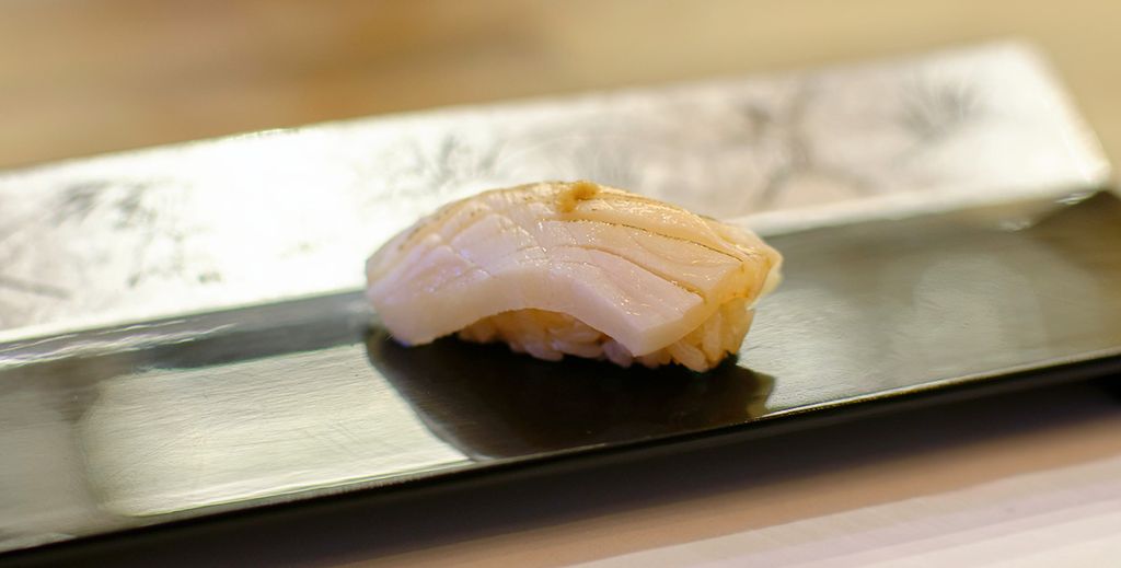 A nigiri sushi prepared with the ingredient escolar lies on a small plate in a sushi restaurant.