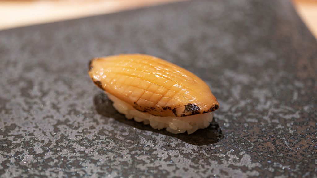 Ein Sushi-Stück mit Awabi, auch bekannt als Abalone (Haliotis), auf einem Reisbett, präsentiert auf einem schwarzen Teller mit gesprenkeltem Muster, ein Beispiel für die traditionelle japanische Küche.