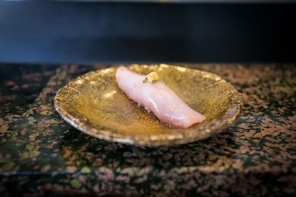 A single piece of albacore sushi nigiri, artfully placed on a golden ceramic plate atop a marble surface.