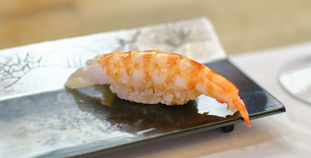 A piece of ebi nigiri sushi, featuring a peeled shrimp atop a small mound of rice, presented on a rectangular ceramic plate with subtle patterning in the background.
