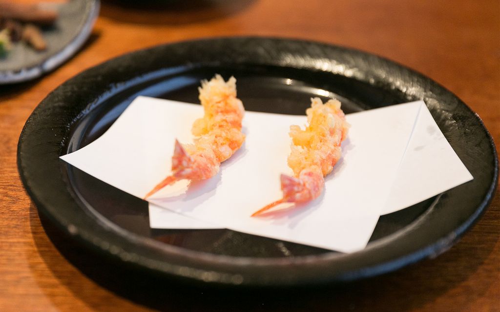 Two pieces of arranged shrimp tempura on white paper placed on a black plate at Mikawa Zezankyo, a Michelin-starred tempura restaurant.
