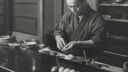 Black-and-white photograph. A man in dark cotton work clothing at a polished wooden counter, hands cupped in the gesture of forming nigiri. Small dishes, a brush, and a glass sit on the counter; a few finished pieces of nigiri are visible at the front edge. Traditional Japanese interior in the background.