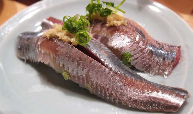 Two pieces of iwashi sardine sushi garnished with ginger and chopped scallions on a white plate