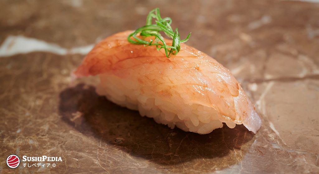 A piece of nigiri sushi, with the ingredient barracuda (kamasu in Japanese), lies on a plate in a sushi restaurant.