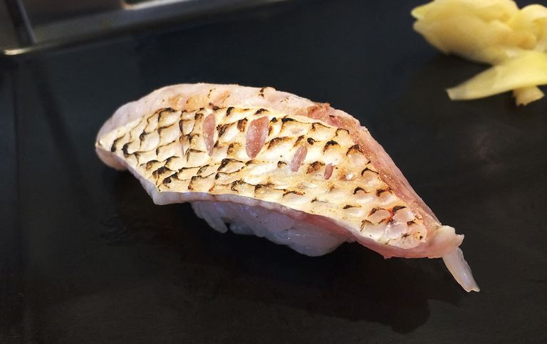 Aburi-style nigiri sushi with grilled fish skin on a black ceramic plate, accompanied by pickled ginger on the side
