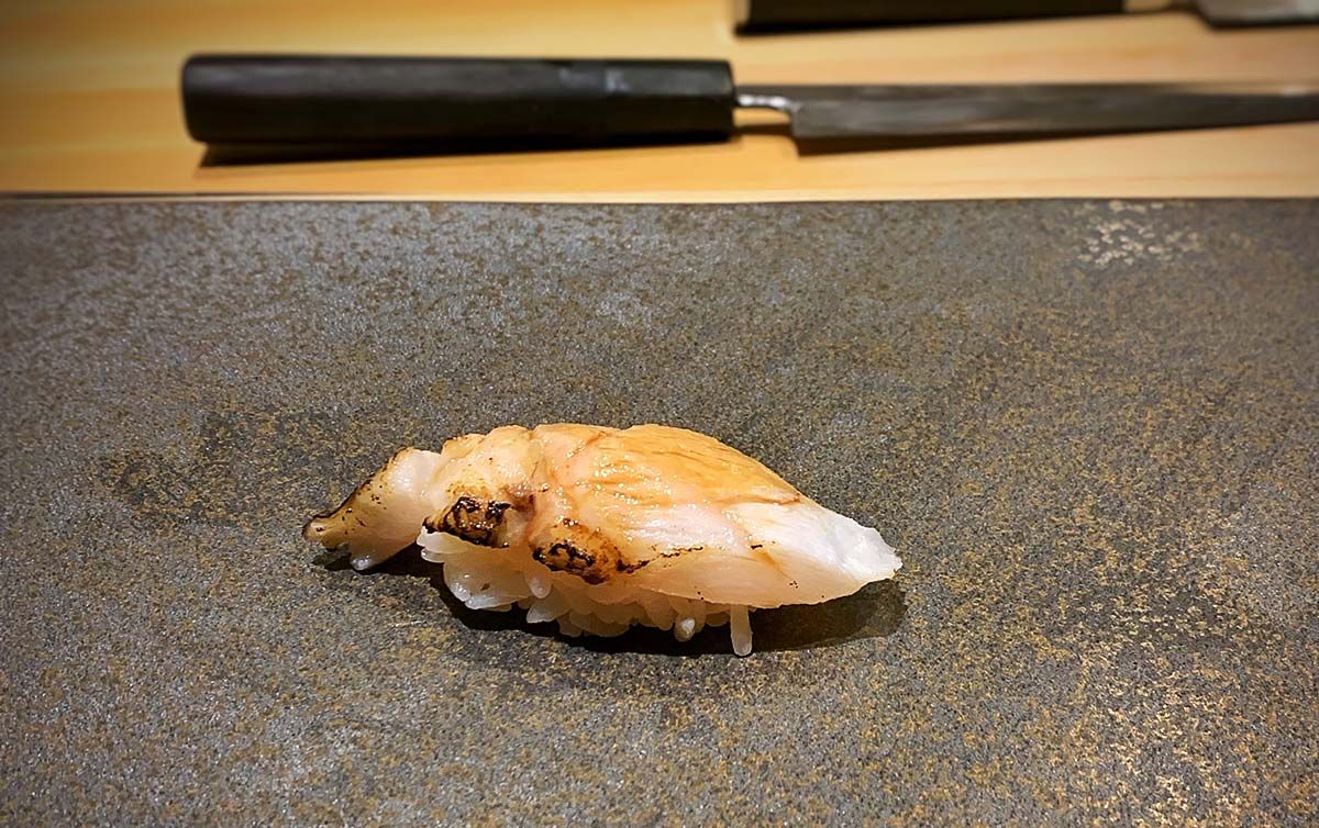Single nigiri sushi with lightly charred nodoguro topping on a dark stone slab table, with blurred traditional Japanese knife in the background