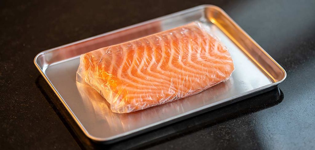 A piece of fresh salmon fillet lying on a metal tray, wrapped in dehydration film. This film removes moisture to preserve the freshness of the salmon, making it ideal for preparing high-quality sushi.