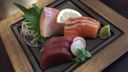 Sashimi on a black plate with raw tuna, salmon, and white fish, served with wasabi, shiso, daikon, and a lemon slice.