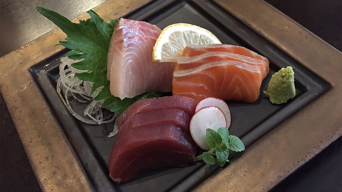 Sashimi on a black plate with raw tuna, salmon, and white fish, served with wasabi, shiso, daikon, and a lemon slice.