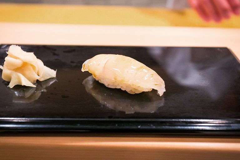 Image displays a single piece of halibut sushi nigiri on a glossy black plate with pickled ginger on the side. The sushi rice is just visible beneath the translucent slice of fish, highlighting the skillful balance of the sushi chef's artistry.