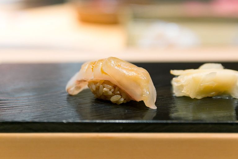 An image showcasing a single piece of halibut nigiri sushi, with a thin, translucent slice of halibut draped elegantly over a mound of vinegared rice, served on a dark, minimalist sushi plate with a blurred background highlighting the simplicity and focus on the sushi itself.