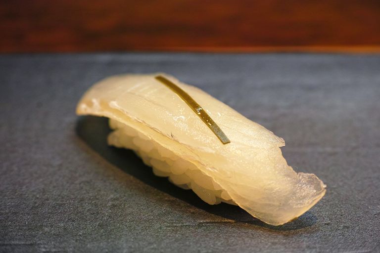 A close-up image of a Hamachi Nigiri sushi. The sushi features a slice of lustrous, pale yellow Hamachi fish, delicately placed over a bed of sushi rice, accented with a thin strip of kelp (kombu) across the top for enhanced umami flavor.