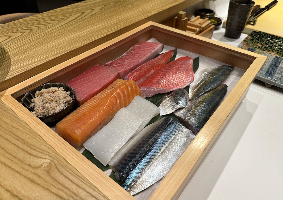 Wooden sushi ingredient box with prepared fish cuts for omakase service, including tuna, salmon, squid, silver-skinned fish and crab meat.