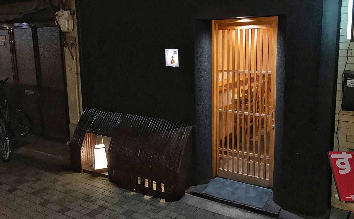 Exterior view of a small sushiya in Osaka at night, with a dark façade, wooden lattice door, and warmly lit entrance.