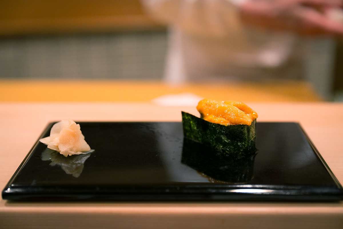 A piece of Uni Gunkan Sushi, consisting of sea urchin roe (uni) on sushi rice, wrapped in a strip of nori seaweed, served on a black plate with pickled ginger beside it.