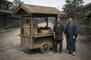 Two men stand beside a wooden street food cart with a thatched roof on a dirt road. The counter holds stacked wooden boxes, boards, and a large round wooden tub (hangiri) used for cooling sushi rice. Traditional wooden buildings and trees line the street in the background, creating a quiet, documentary-style scene.