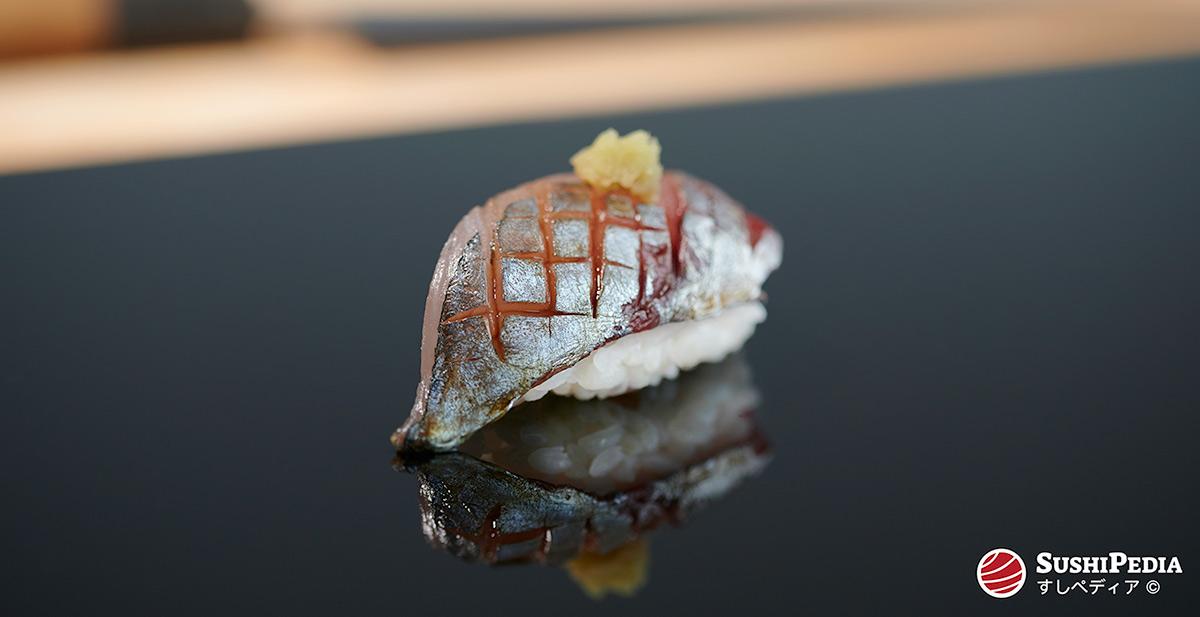A piece of bastard mackerel or horse mackerel nigiri sushi that has been glazed with soy sauce and is sitting on the counter of a sushi restraurant.