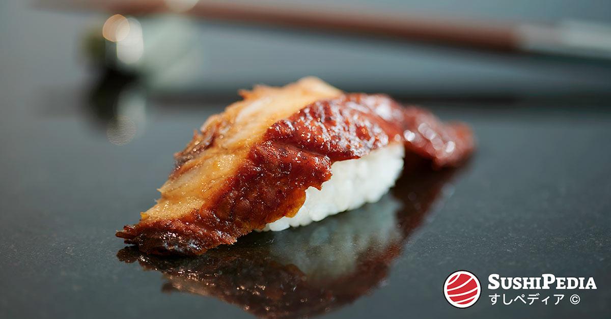 Ein Stück Aal-Sushi (als Nigiri geformt), dass mit Aal-Sauce (unagi no tare) bestrichen wurde und auf der Theke eines Sushi-Restraurants liegt.